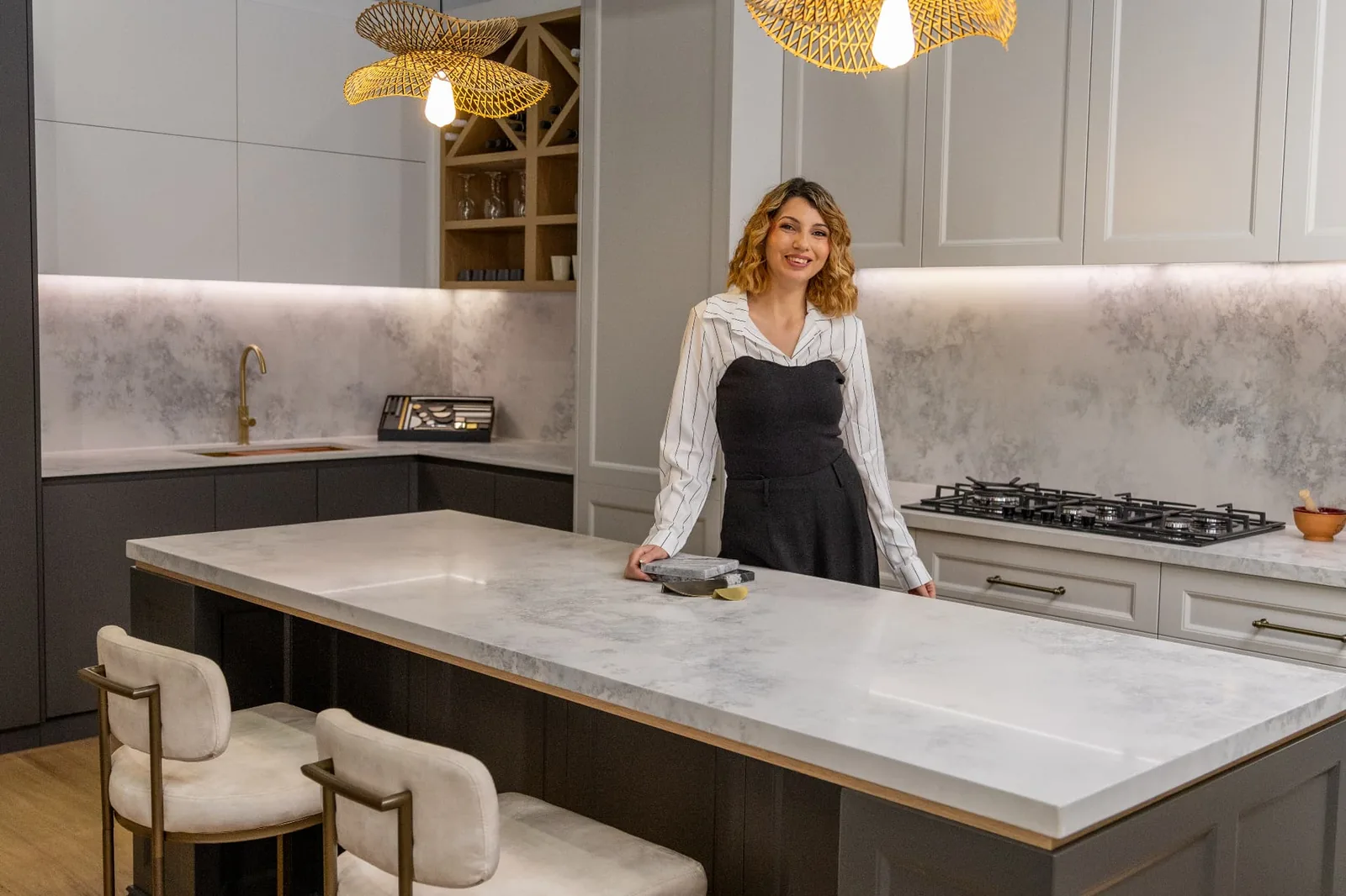 A designer standing in a completed Haute Kitchen with marble-look stone island bench, custom joinery and feature pendant lighting